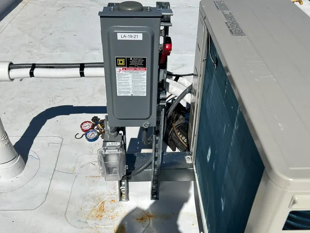 Gray rooftop disconnect switch on a metal strut frame beside an HVAC condensing unit, fed by liquid-tight flexible conduit, commercial building