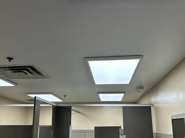 Restroom ceiling with several square LED panel lights turned on above dark stall partitions