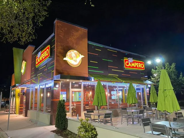 Pollo Campero restaurant in Raleigh, NC at night, with illuminated Pollo Campero signs, a lit glass storefront and a patio with closed green umbrellas
